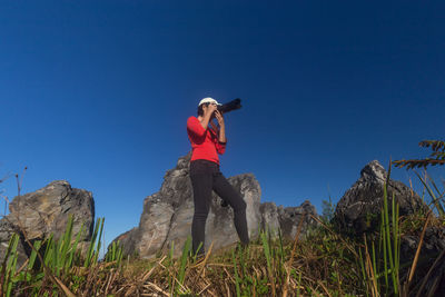 Low angle view of woman photographing through camera while standing on mountain against clear blue sky