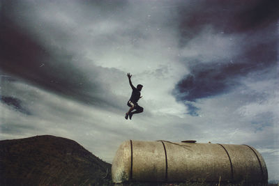 Man jumping on storage tank by mountain against sky
