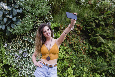Beautiful woman taking selfie from mobile phone while standing against plants at park