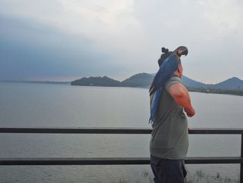 Man with macaw on shoulder standing by lake against cloudy sky
