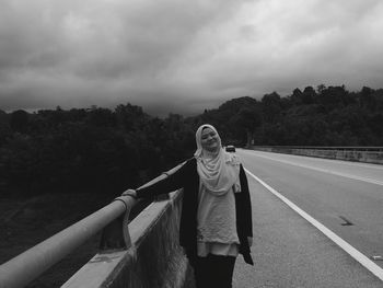 Young woman in hijab standing by railing on bridge against cloudy sky