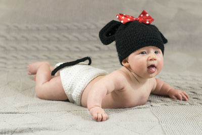 Cute baby wearing knit hat with red bow lying on bed at home