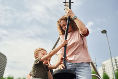 Happy adorable little girl riding rope swing while having fun on playground under supervision of mother