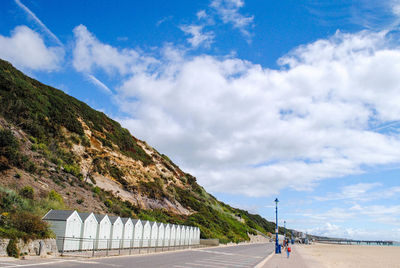 Footpath and beach huts by mountain against cloudy sky