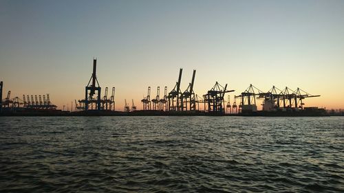 Silhouette cranes at harbor against sky during sunset