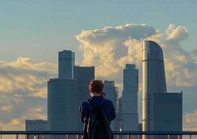 Rear view of man looking at modern buildings in city against cloudy sky