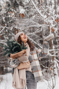 A stylish girl in a snowy forest holds branches of a nobilis fir with a garland, smiles and throws 