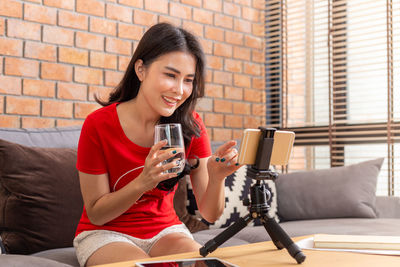 Smiling young woman holding drink sitting against the wall