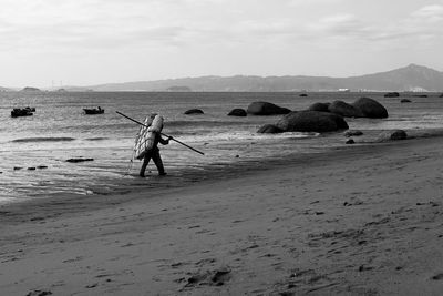 Man carrying luggage while walking on shore at beach against sky