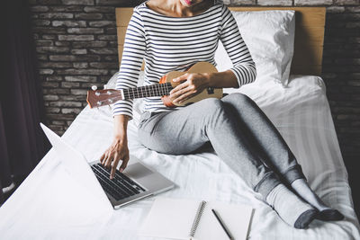 Low section of woman using laptop while playing ukulele on bed at home