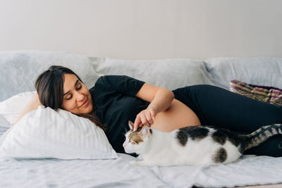 Pregnant middle-aged woman petting a cat while lying on the sofa at home.