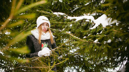 Young woman wearing warm clothing while holding disposable cup amidst trees during winter