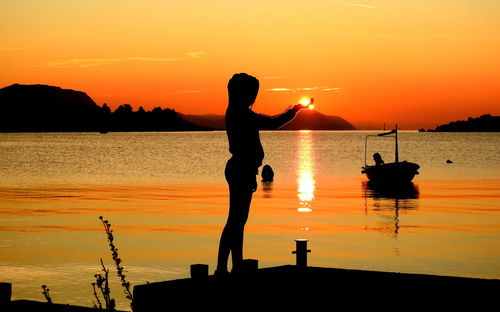 Optical illusion of silhouette woman holding sun at sea shore during sunset