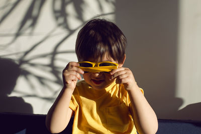 Boy in a yellow t-shirt and sunglasses against a white wall with the shadow of a palm tree