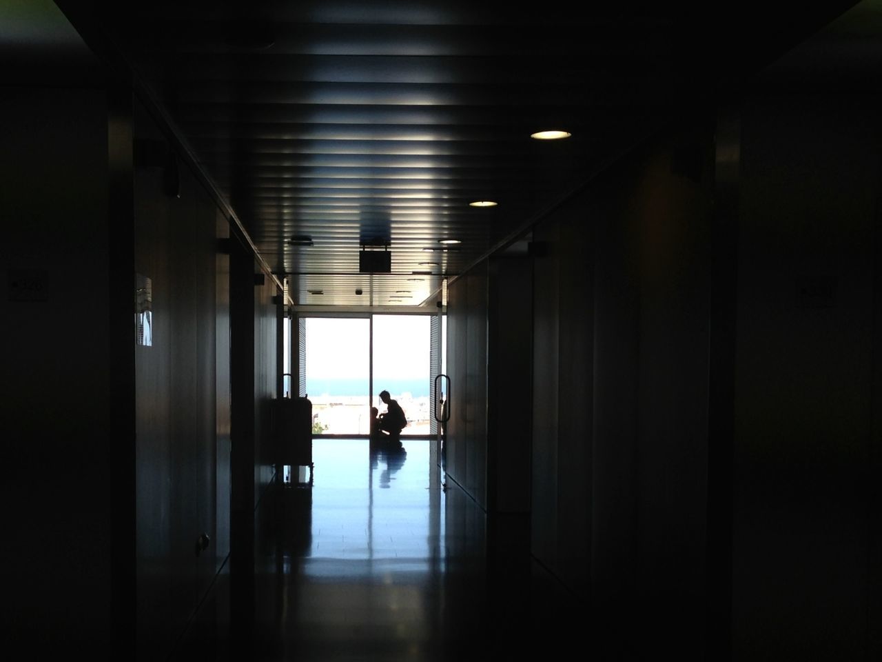 indoors, ceiling, corridor, illuminated, architecture, silhouette, built structure, window, lighting equipment, full length, reflection, flooring, walking, door, glass - material, rear view, wall, the way forward