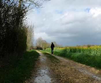 Rear view of man walking on dirt road by field against cloudy sky