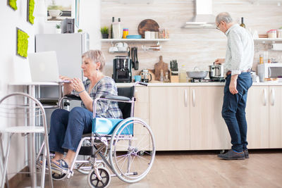 Disabled woman on wheelchair using laptop with man in background at home