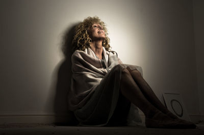 Smiling mature woman wrapped in blanket sitting on floor against wall at home