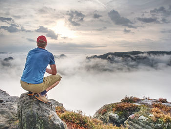 Man sitting on rock looking at waterfall against sky