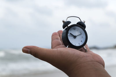 Cropped hand of person holding alarm clock against sky