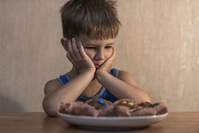 Thoughtful boy with cupcakes sitting at table against wall