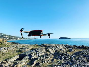 Drone flying on rocks by sea against clear blue sky