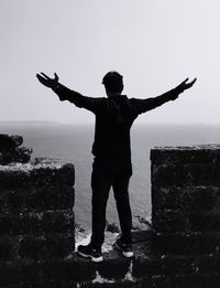 Full length of man with arms outstretched standing on retaining wall against sea and clear sky