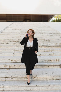 Content female manager in elegant suit walking down the stairs in city and discussing work issues on cellphone while looking away