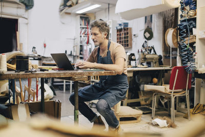 Male upholstery worker using laptop at workbench in workshop