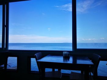 Chairs and table by sea against sky seen through window