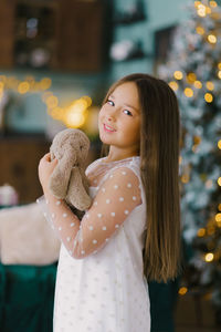 The kid near the christmas tree holds a plush bunny with a soft toy. the girl is happy