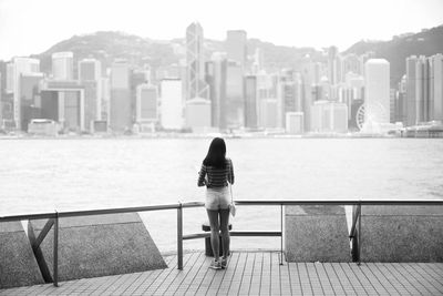 Rear view of young woman standing on promenade by sea against city