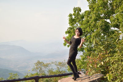 Portrait of smiling woman gesturing horn sign while standing on rock against mountains