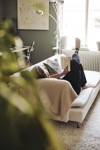 Mature woman reading book while lying down on sofa at home