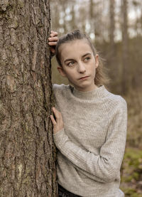 Thoughtful teenage girl looking away while standing behind tree in forest