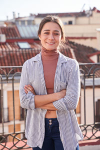 Cheerful young female in casual outfit standing with crossed arms on terrace and looking at camera