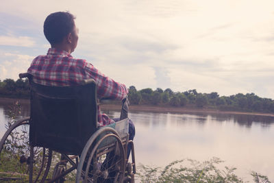 Rear view of man sitting by lake against sky