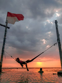 Man resting on hammock over sea against cloudy sky during sunset