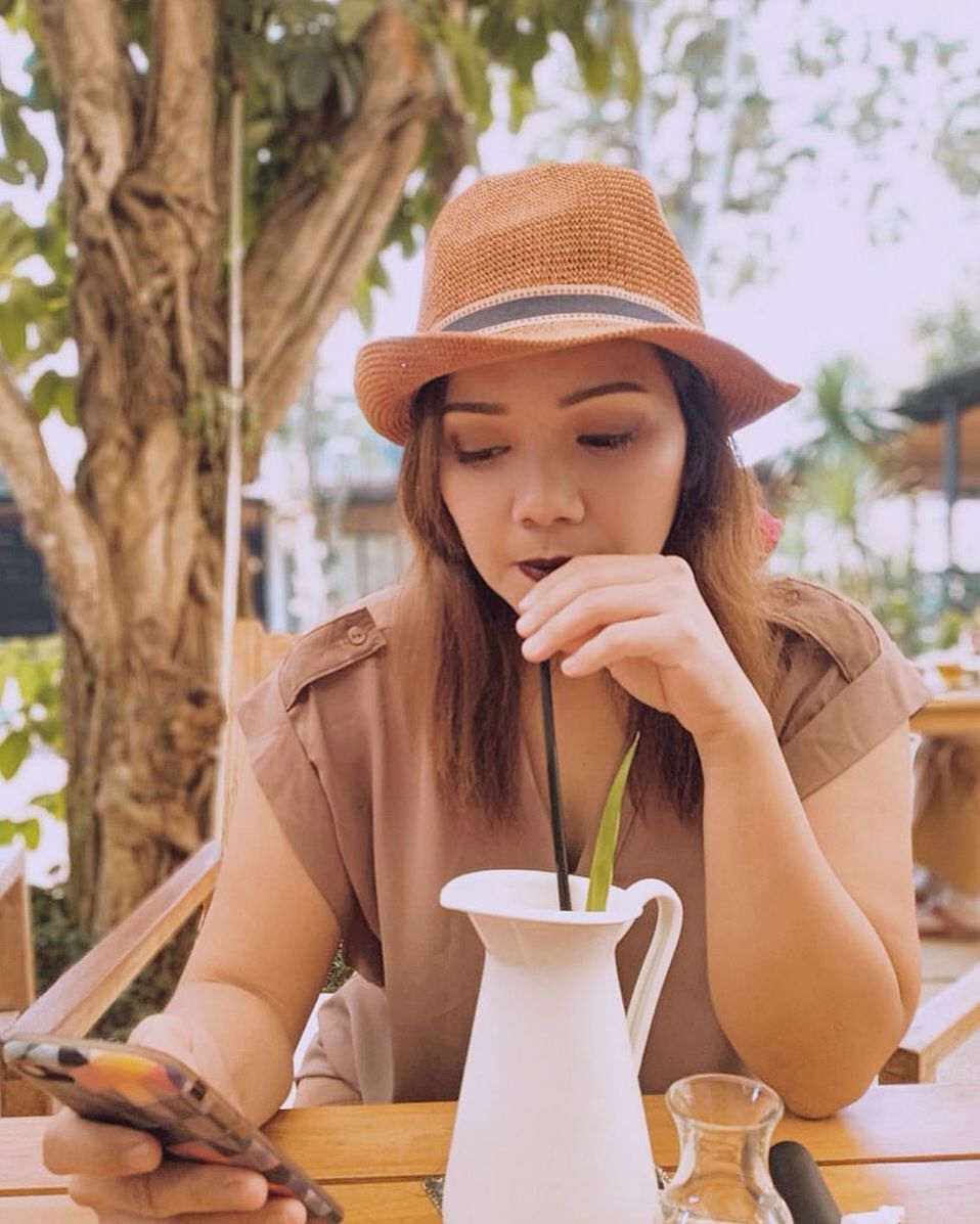 hat, leisure activity, drink, lifestyles, food and drink, clothing, front view, one person, sitting, young adult, real people, young women, cup, table, refreshment, coffee - drink, drinking, portrait, holding, drinking straw, outdoors, contemplation