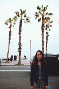 Portrait of young woman standing by palm trees against sky