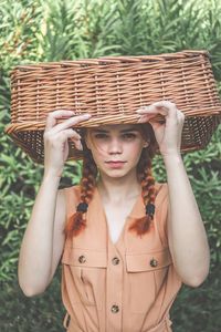 Portrait of young woman holding basket on head while standing against plants