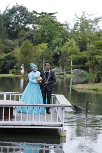 Portrait of bride and bridegroom standing on pier over lake against trees