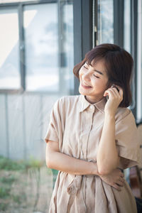 Smiling young woman listening music while standing by window at home