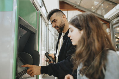 Father using atm machine while standing with daughter at railroad station