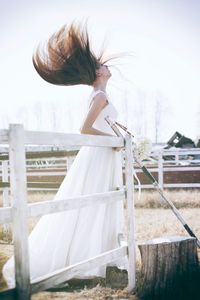 Beautiful bride with tousled hair standing by railing in pen against clear sky