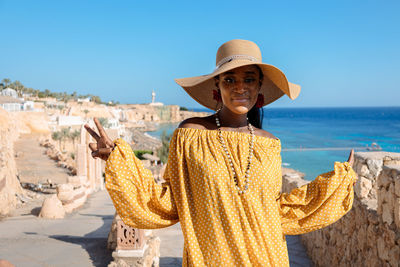 Happy african american woman in yellow dress and sun hat enjoys view of coast of red sea on natural 