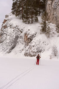 Rear view of young man in red brown clothes with backpack skiing near rocks  winter sports hiking