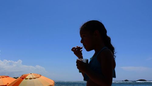 Side view of girl eating food against blue sky on sunny day