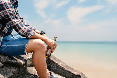 Side view midsection of man holding beer bottle while sitting on rock at beach