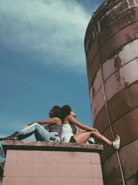 Low angle view of female friends sitting on built structure against blue sky